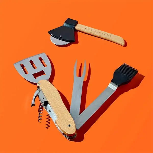 A set of barbecue tools including a pizza cutter axe, spatula, fork, corkscrew and basting brush with wooden handles, arranged on a bright orange background.